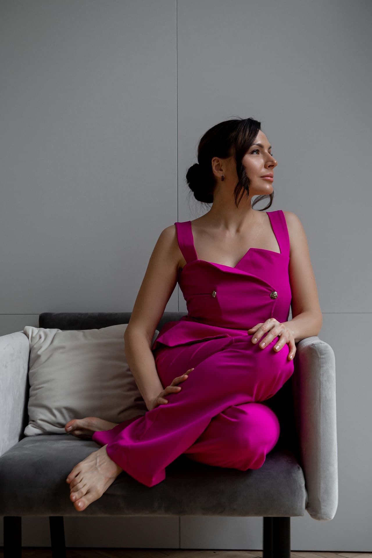 Woman in a vibrant pink outfit sits barefoot on a grey armchair, looking away.